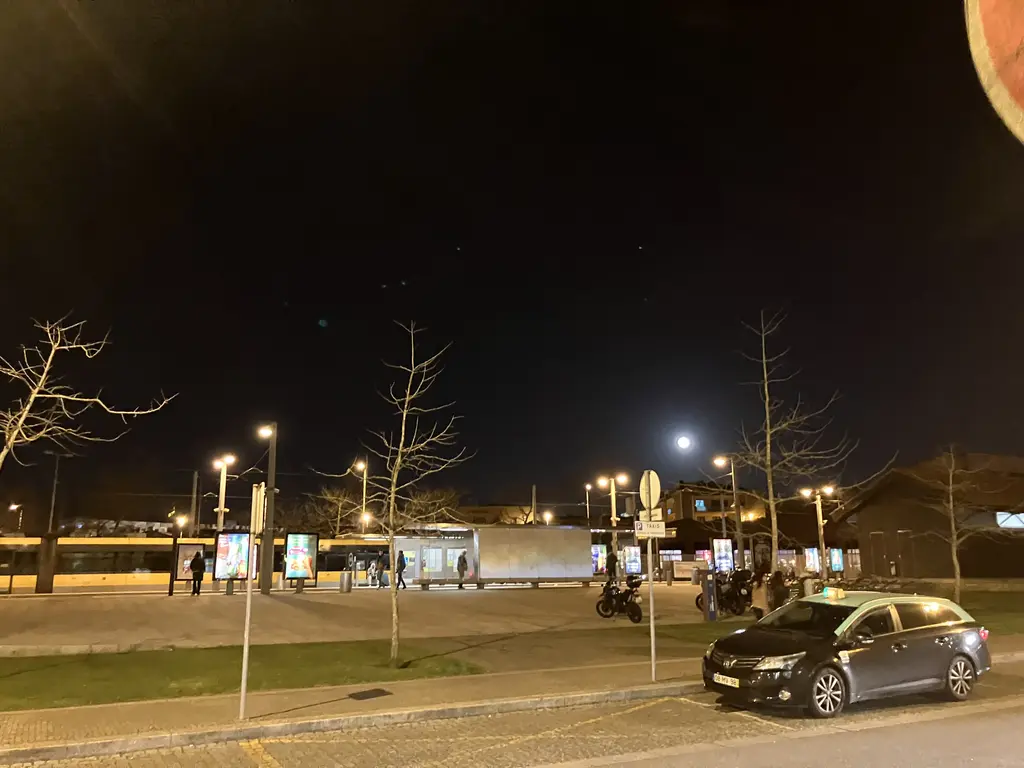 La estación de metro de Póvoa de Varzim, con luces encendidas y pasajeros esperando. La luna llena brilla en el cielo despejado, reflejando la tranquilidad de la escena urbana.