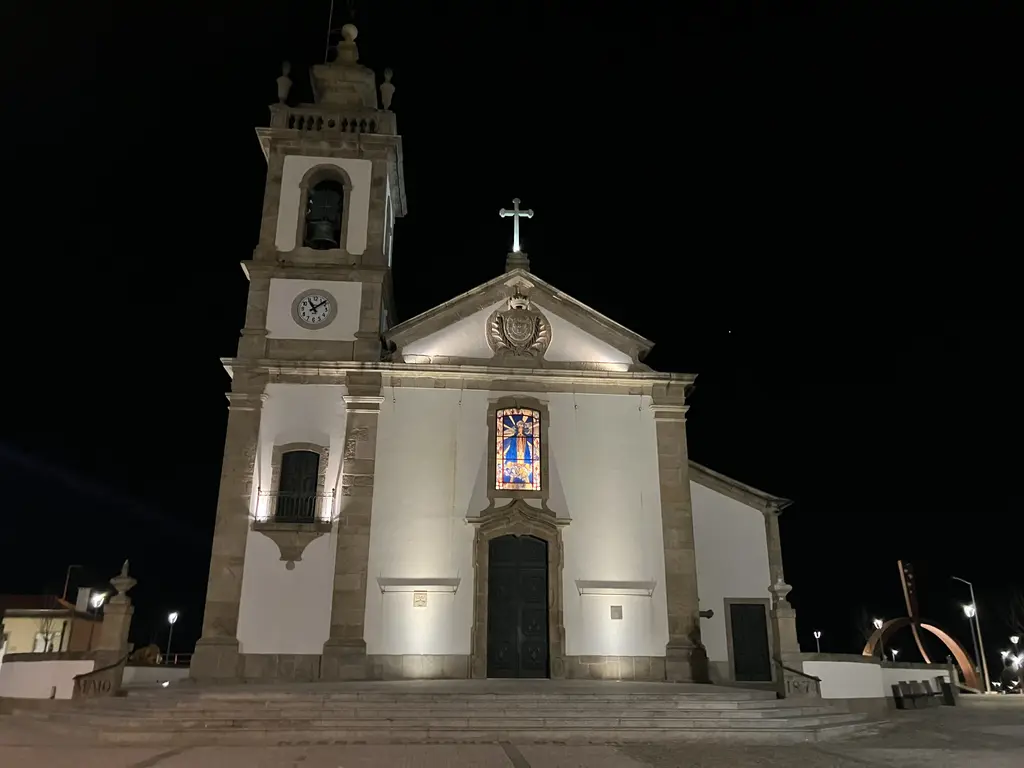 Fotografía nocturna de la Iglesia Matriz de Póvoa de Varzim, iluminada y destacando su torre con reloj, su fachada de piedra y su vitral colorido en la entrada.