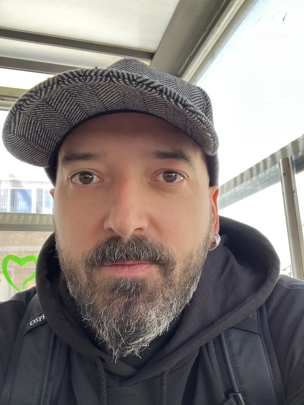 Primer plano de un hombre con barba canosa, gorra y sudadera negra, tomándose una foto bajo una estructura metálica con cristales y un grafiti de un corazón verde.
