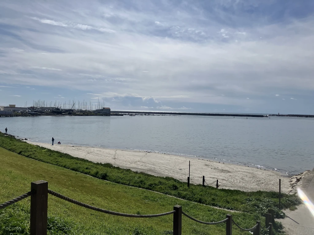 Vista de una pequeña playa de arena con césped en primer plano, el mar tranquilo y un puerto con barcos al fondo. Unas pocas personas disfrutan del lugar bajo un cielo parcialmente nublado.