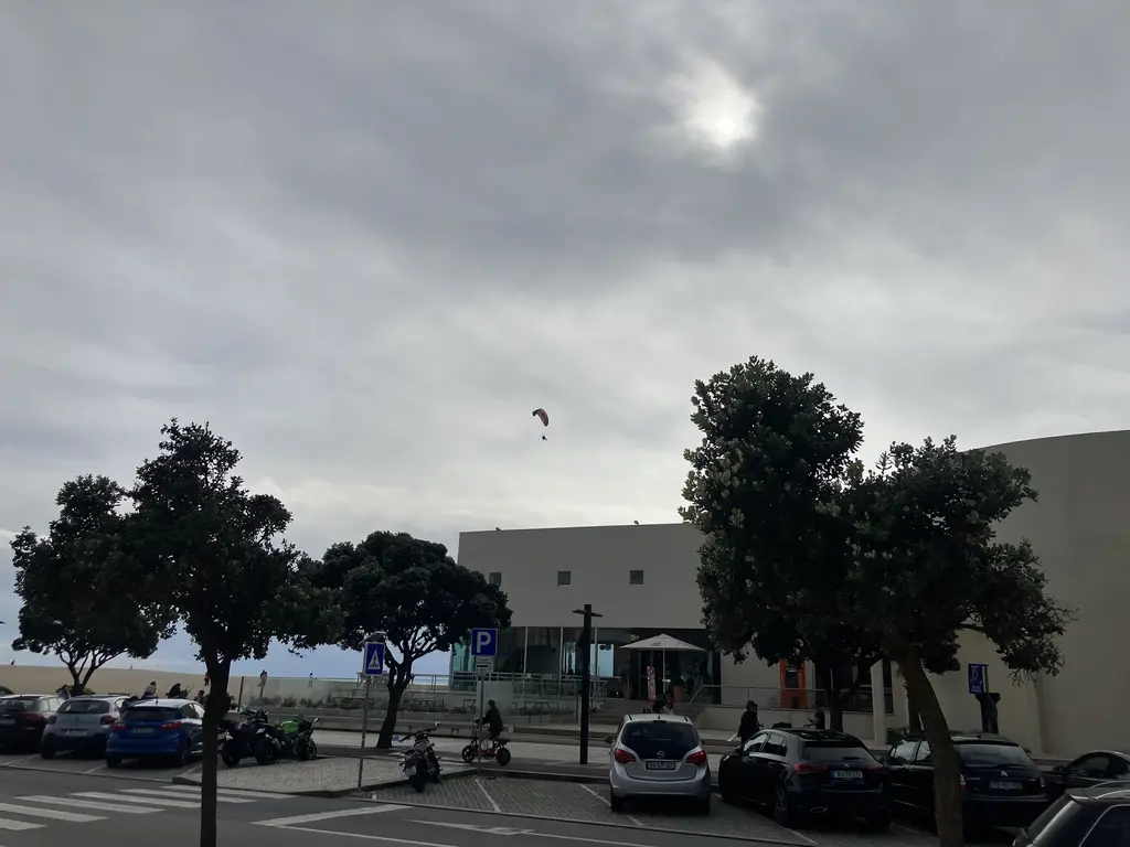 Un parapente volando sobre la zona costera de Póvoa de Varzim en un día nublado. En la parte inferior, se ven árboles, coches estacionados y un edificio blanco moderno.