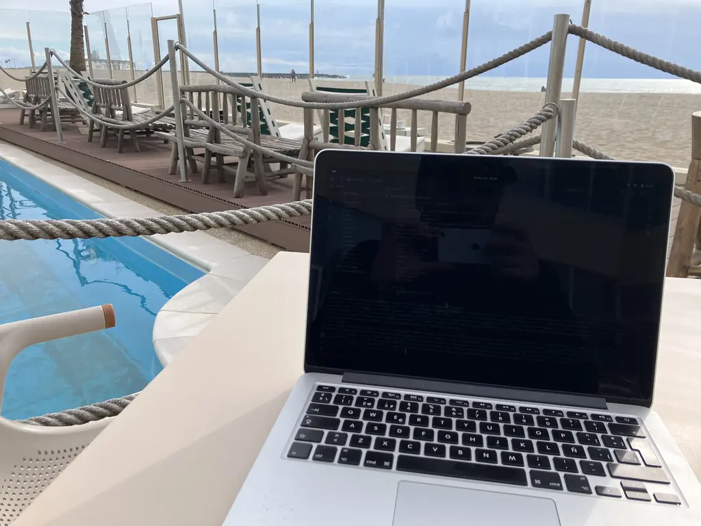 Un ordenador portátil sobre una mesa junto a una piscina, con el reflejo del fotógrafo en la pantalla. De fondo, la arena, el mar y un cielo parcialmente nublado.