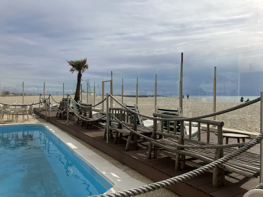 Zona de descanso junto a una piscina con sillas de madera y cuerdas de separación, con una vista directa a la playa y el océano bajo un cielo nublado.