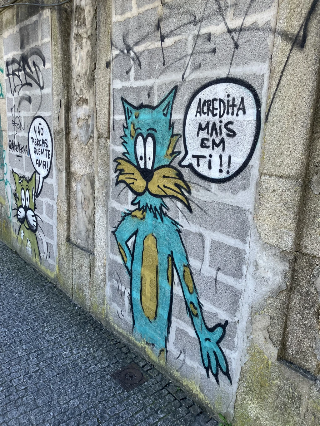 Mural en una pared de ladrillos con un dibujo de un gato azul con manchas marrones, que dice "Acredita mais em ti!!" ("Cree más en ti!!") en un bocadillo. A la izquierda, otro mensaje en portugués anima a no perder lo que se ama.
