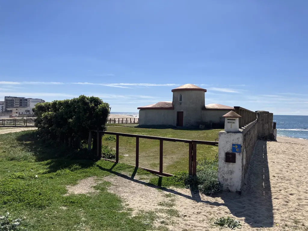 Antigua capilla con muros blancos y techo de tejas rojas, rodeada por una valla de madera. A su derecha, un camino de arena que lleva al mar, con un cielo despejado.