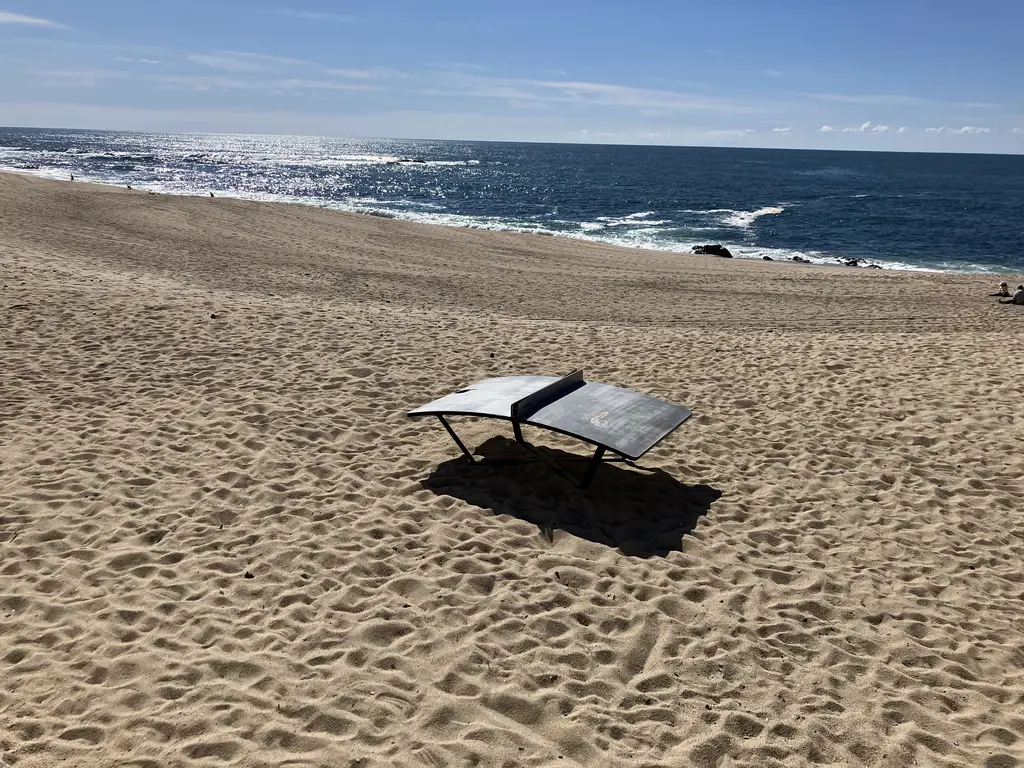 Una mesa de Teqball sobre la arena de la playa, diseñada con una superficie curva para jugar con una pelota de fútbol. Al fondo, el mar azul brilla con el reflejo del sol y el cielo despejado.