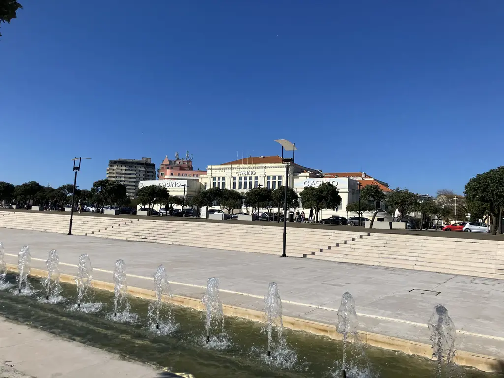 Fuente con chorros de agua alineados en una plaza de Póvoa de Varzim. De fondo, el casino y edificios con arquitectura clásica bajo un cielo azul despejado.