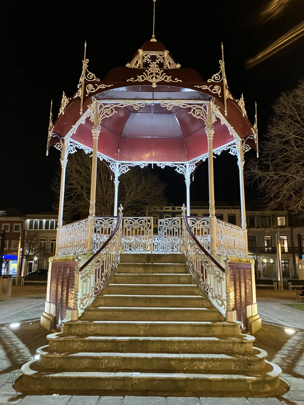 Imagen de un quiosco ornamental con techo rojo y estructura de hierro forjado iluminado en la noche. Se encuentra en una plaza de Póvoa de Varzim, Portugal, con un fondo de edificios históricos y árboles sin hojas.