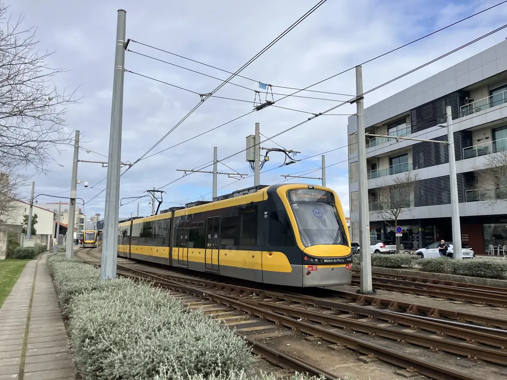 Metro do Porto circulando por vías entre arbustos y edificios modernos. El cielo nublado contrasta con la vibrante escena urbana.