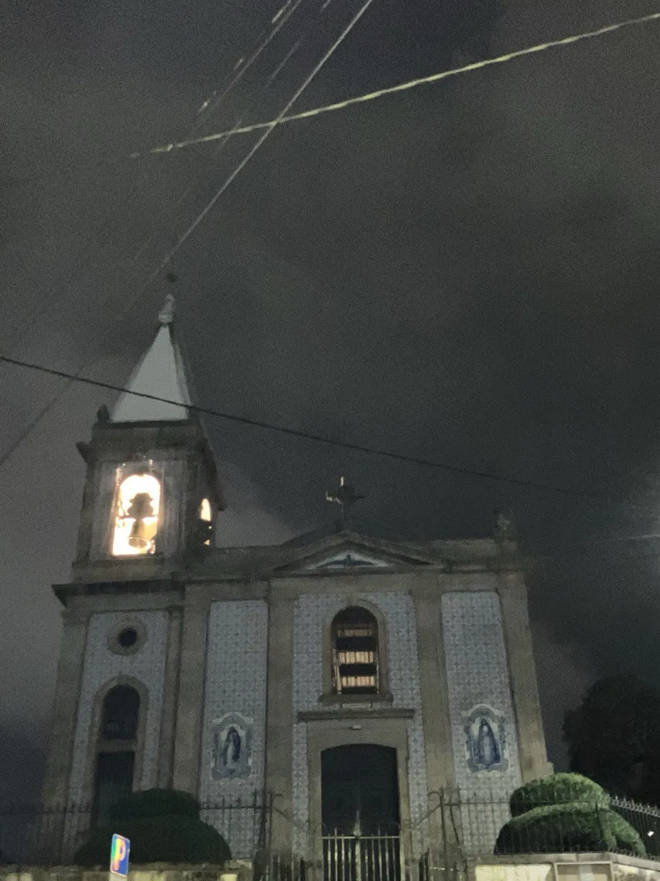 Fachada iluminada de noche de otra iglesia en Vila Nova de Gaia, con revestimiento de azulejos, torre campanario con campana visible, cruz en la cumbre y arbustos recortados en primer plano.