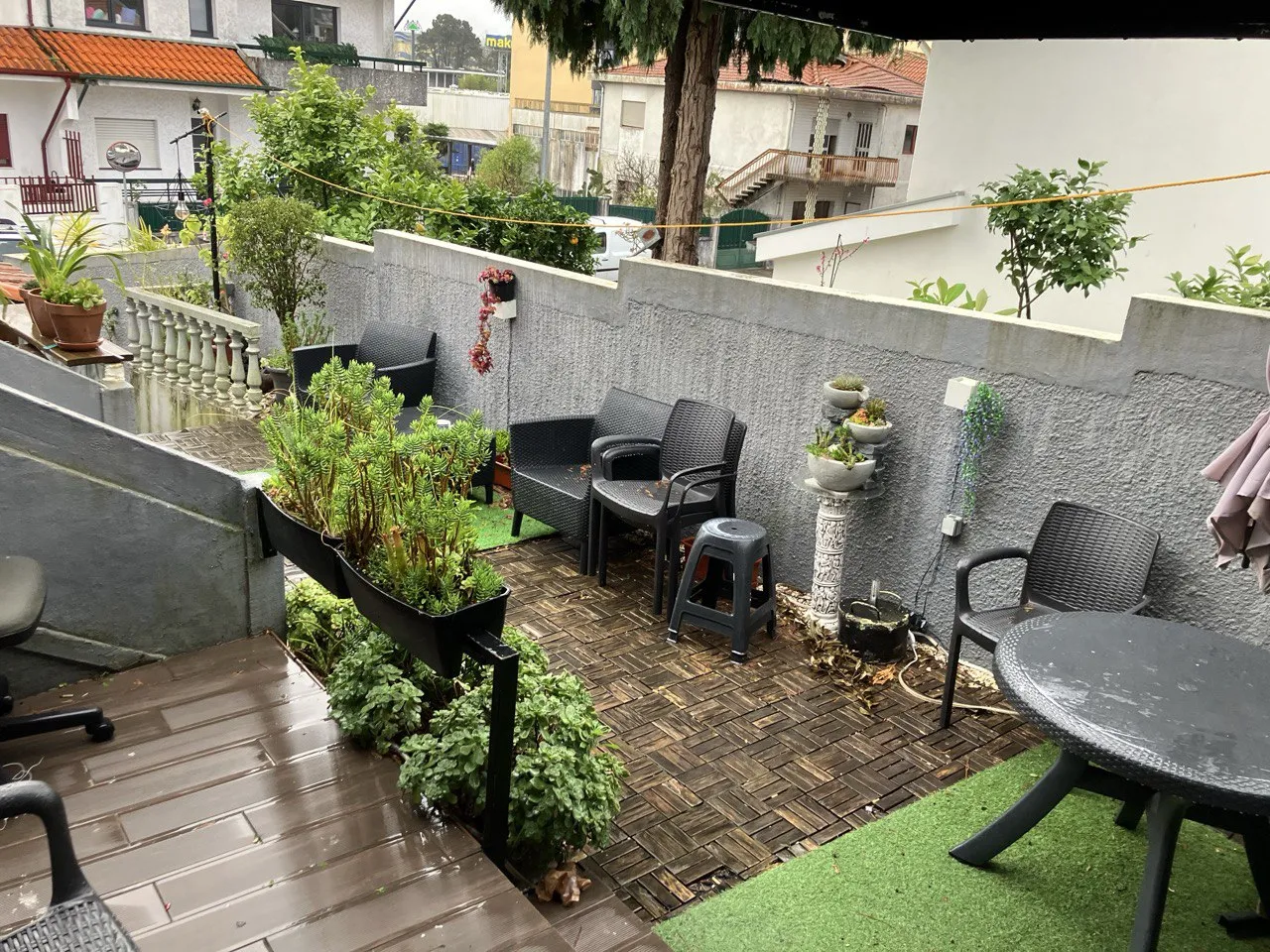 Terraza exterior diurna en Vila Nova de Gaia con suelo de tarima y césped artificial, varias macetas con plantas suculentas, sillas y mesa de plástico negro junto a un muro gris bajo un tendedero.