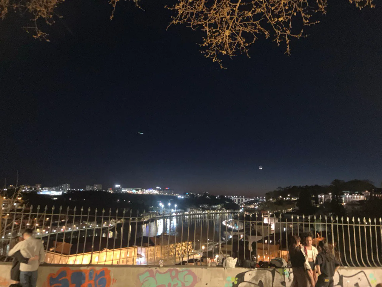 Vista nocturna del río Douro desde un mirador en Oporto, con luces de la ciudad reflejadas en el agua, una luna en cuarto creciente en el cielo, y varias personas conversando junto a una baranda con grafitis.