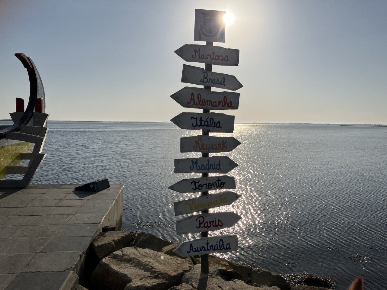 Cartel de direcciones ubicado junto a un muelle con vista al mar, señalando diferentes destinos como Brasil, Alemania, Italia, Madrid, Toronto, Venezuela y París. El sol brilla en el horizonte, reflejándose en el agua, mientras una estructura de madera con forma de barco se encuentra a la izquierda. La escena transmite una sensación de aventura, exploración y posibilidades infinitas.