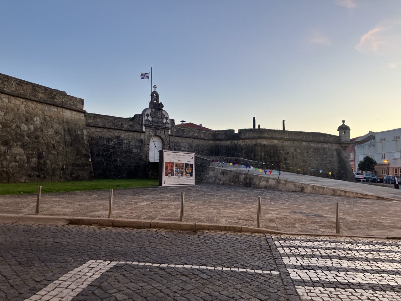 Vista exterior de una fortaleza de piedra en Póvoa de Varzim, con muros altos, una bandera ondeando en la parte superior y una entrada principal flanqueada por carteles, sobre un pavimento empedrado al atardecer.