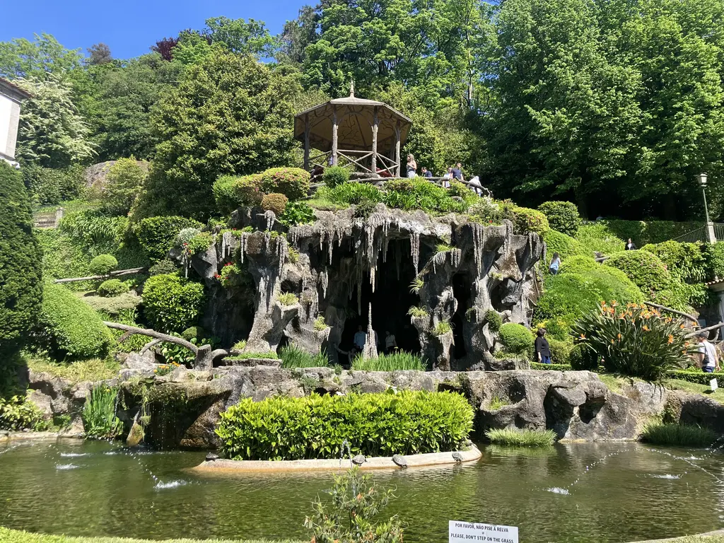 Jardín con una gruta decorativa y una cascada artificial, rodeado de vegetación perfectamente podada y visitantes disfrutando del lugar.