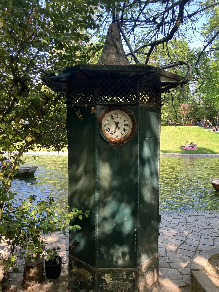 Reloj antiguo montado en una estructura de hierro verde junto a un lago, con una barca y personas descansando en un parque.