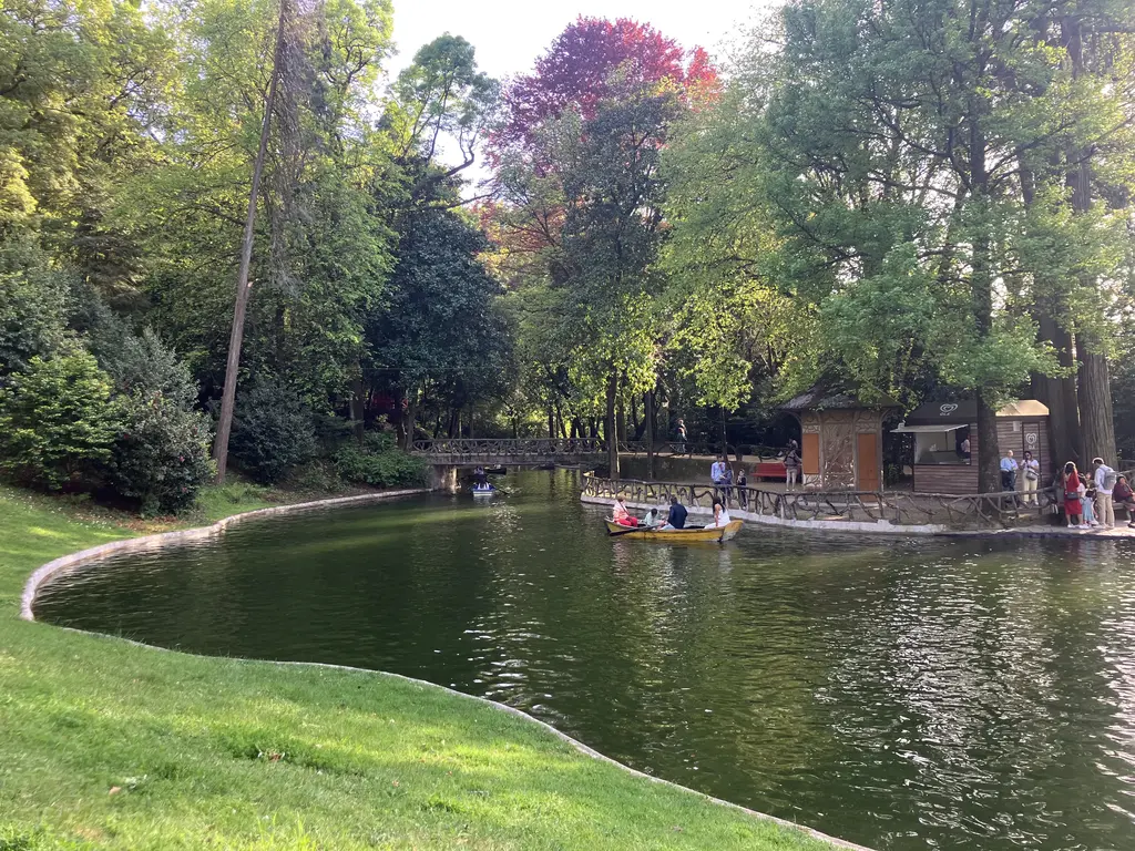Lago rodeado de árboles y vegetación en un parque, con personas paseando en bote y una caseta de madera junto al muelle.