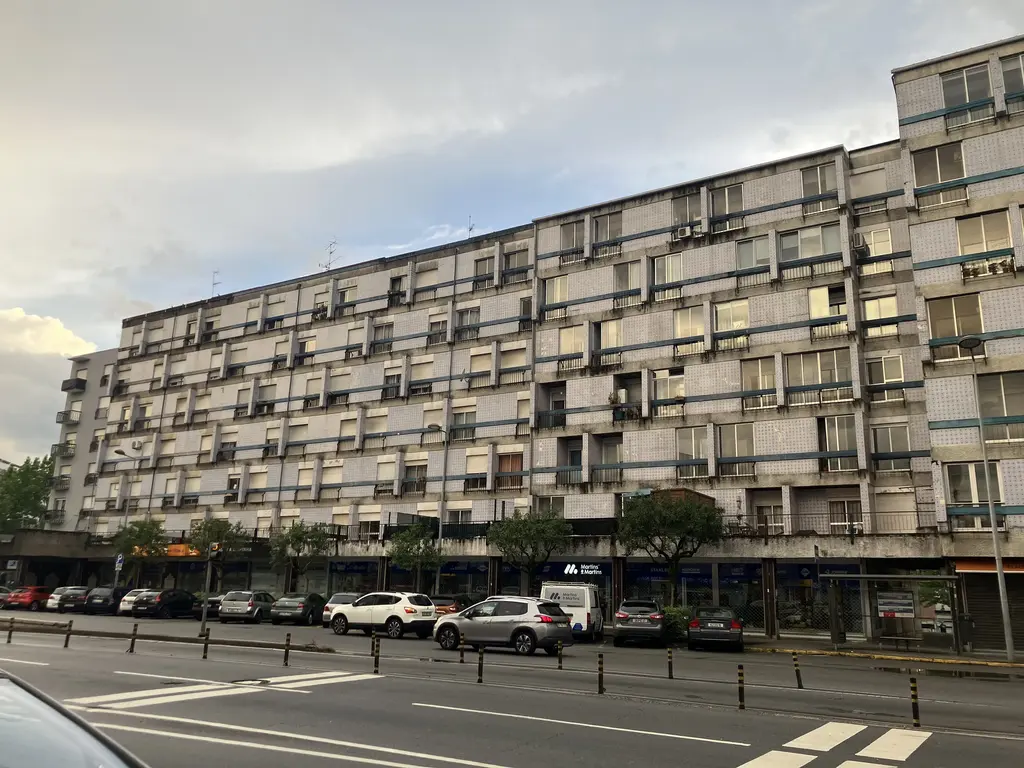 Fachada de un edificio residencial largo y desgastado, con pequeños balcones cerrados y tiendas en la planta baja, al atardecer.