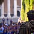 César Emperador de Roma pronunciando un discurso al pueblo romano en una plaza abarrotada frente a su palacio visto desde lejos.